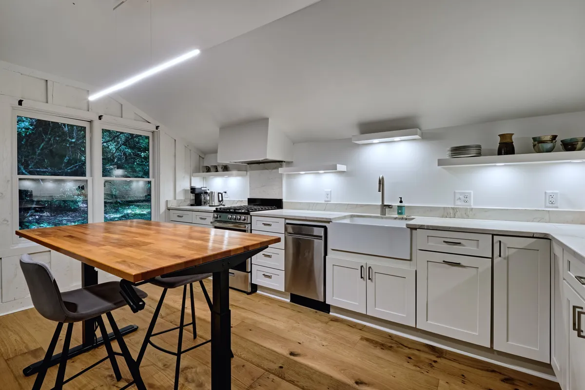 The image features a stylish dining area set next to a kitchen with stainless steel appliances. A wooden dining table is positioned beside large windows, offering lovely woodland views and ample natural light. The scene feels both contemporary and cozy, designed for intimate meals or relaxed family gatherings.
