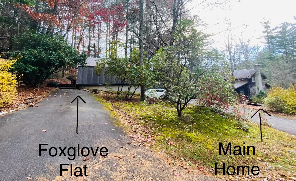 A wider exterior view showing Foxglove Flat nestled amidst mature trees, with glimpses of the main home and a meandering driveway. The setting emphasizes the cottage’s seclusion while also subtly connecting it to the nearby community. The natural landscaping and open spaces communicate both serenity and a sense of welcome.