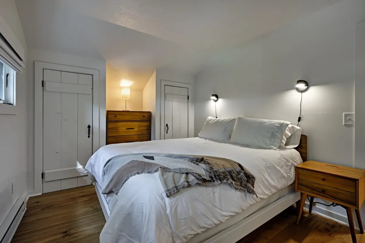 This image features a serene bedroom with a bed dressed in white plush bedding, complemented by wooden nightstands and a decorative throw blanket. Soft, natural light streams through nearby windows, and wall-mounted reading lights add to the tranquil ambiance. The décor is simple and calming, reinforcing a restful atmosphere.