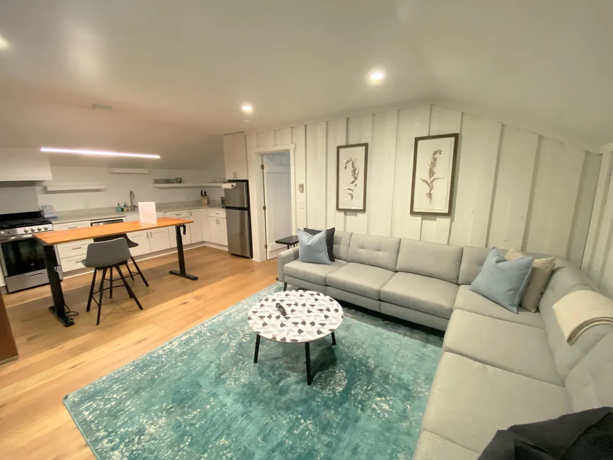 A bright, welcoming living space with a sectional sofa, coffee table, and a view into the kitchen island. The room feels lively and social, suggesting moments of relaxation, group conversation, or family entertainment, and offers a broader perspective on the central living area.