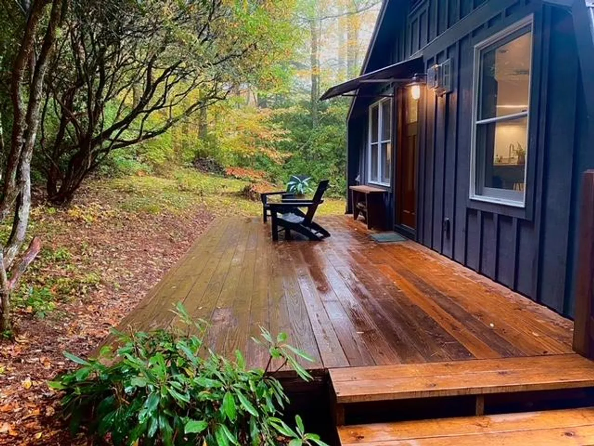This image features a charming wooden deck attached to the cottage, with an adjoining chair perfectly positioned to soak in the surrounding lush trees. The doorway hints at the transition between indoor comfort and the embrace of nature. The tranquil scene invites relaxation, reading, or simply enjoying the peaceful woodland environment.